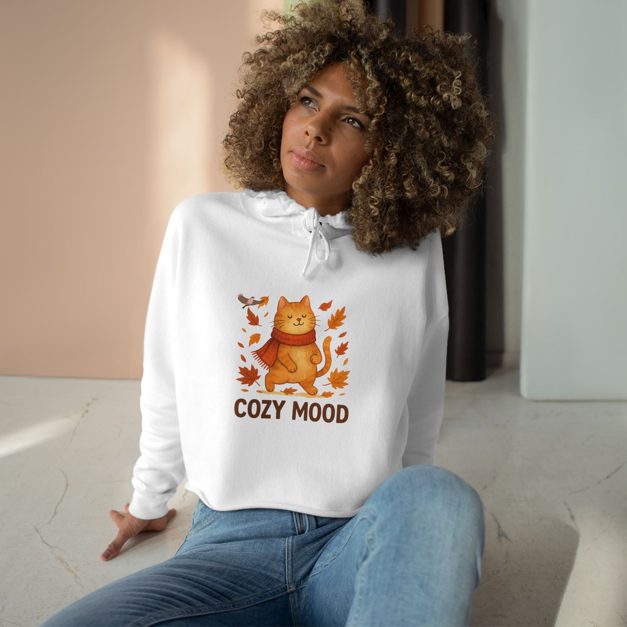 Woman wearing a white hoodie with a cat graphic and 'Cozy Mood' text, sitting indoors.