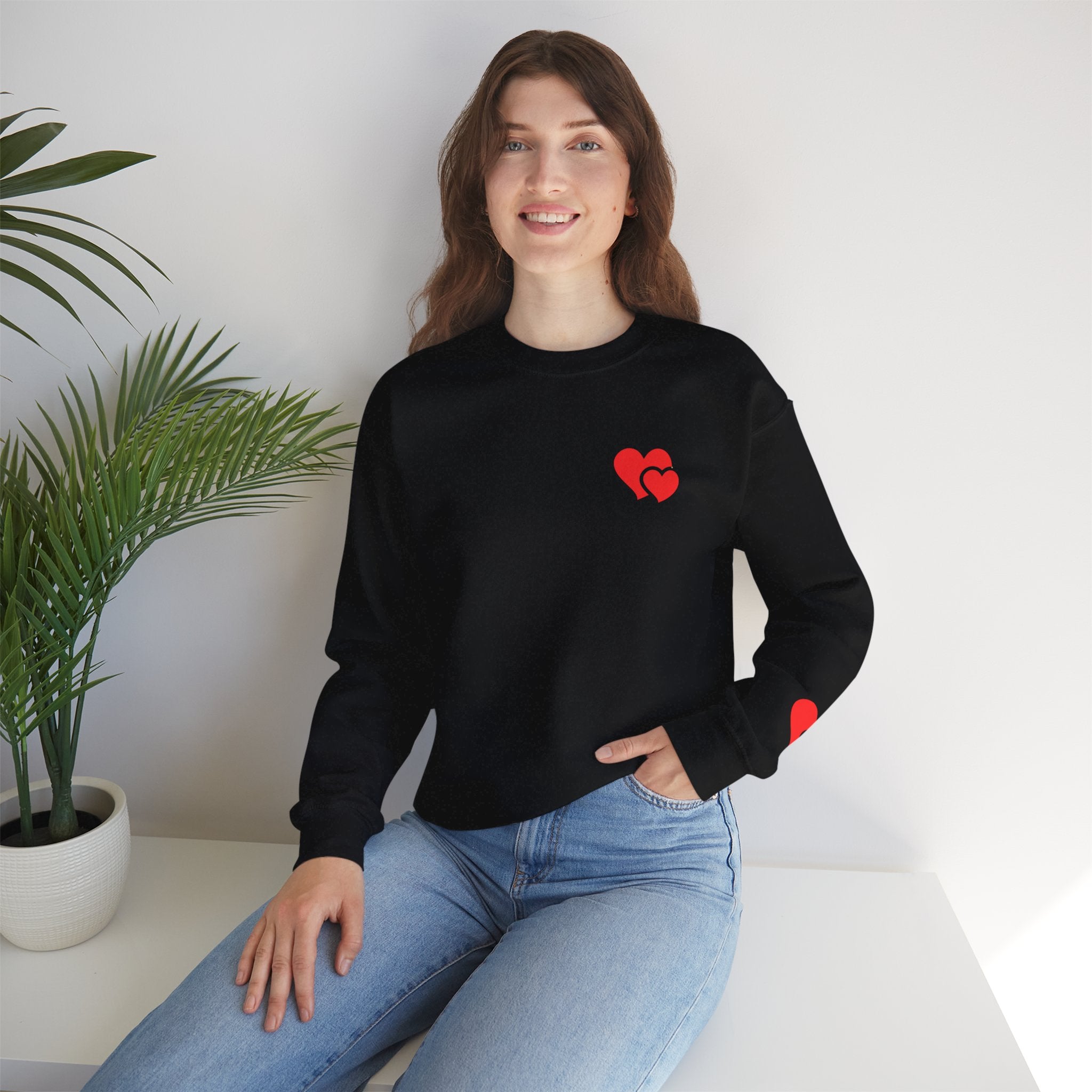 Woman wearing a black sweatshirt with a red heart design, sitting on a white surface with a plant in the background.