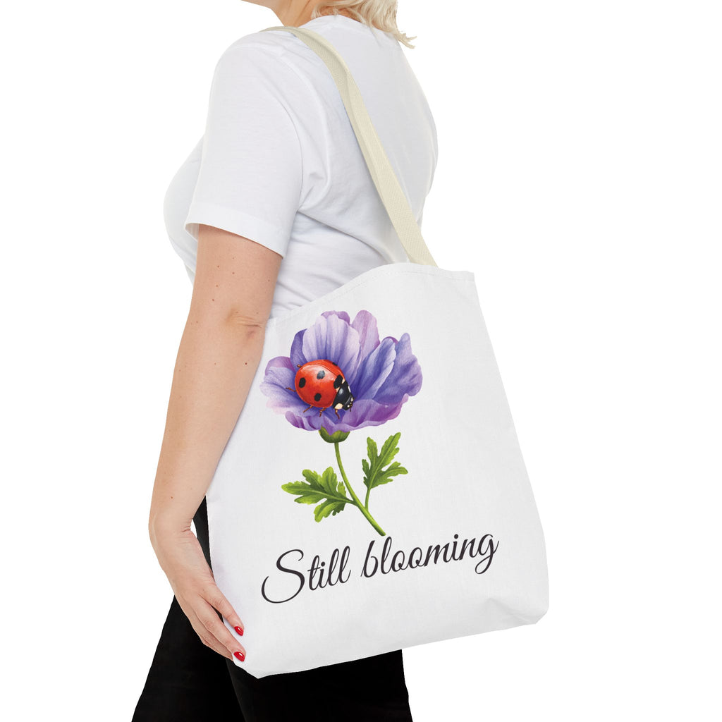 Person holding a tote bag with a purple flower and ladybug design, and the text 'Still Blooming'.