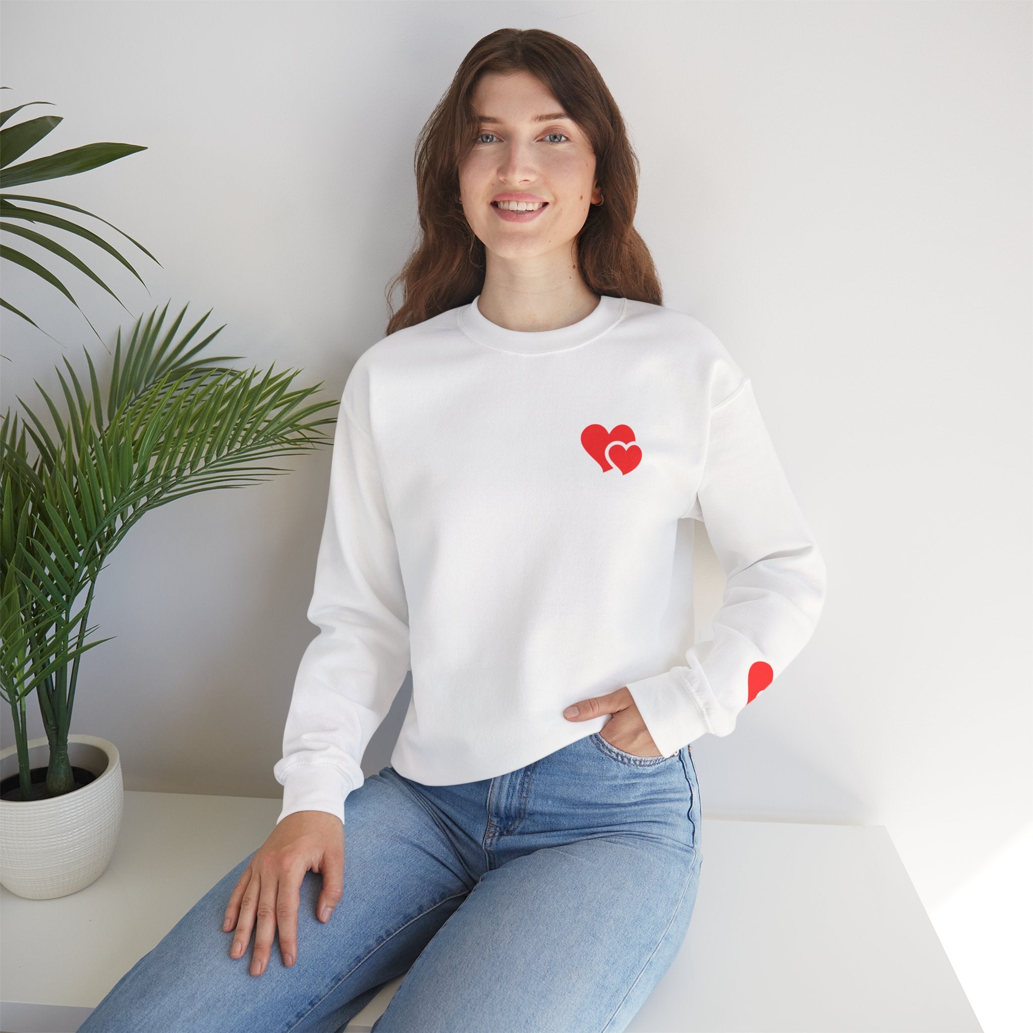 Woman wearing a white sweatshirt with red heart designs sitting on a white surface with a plant in the background.