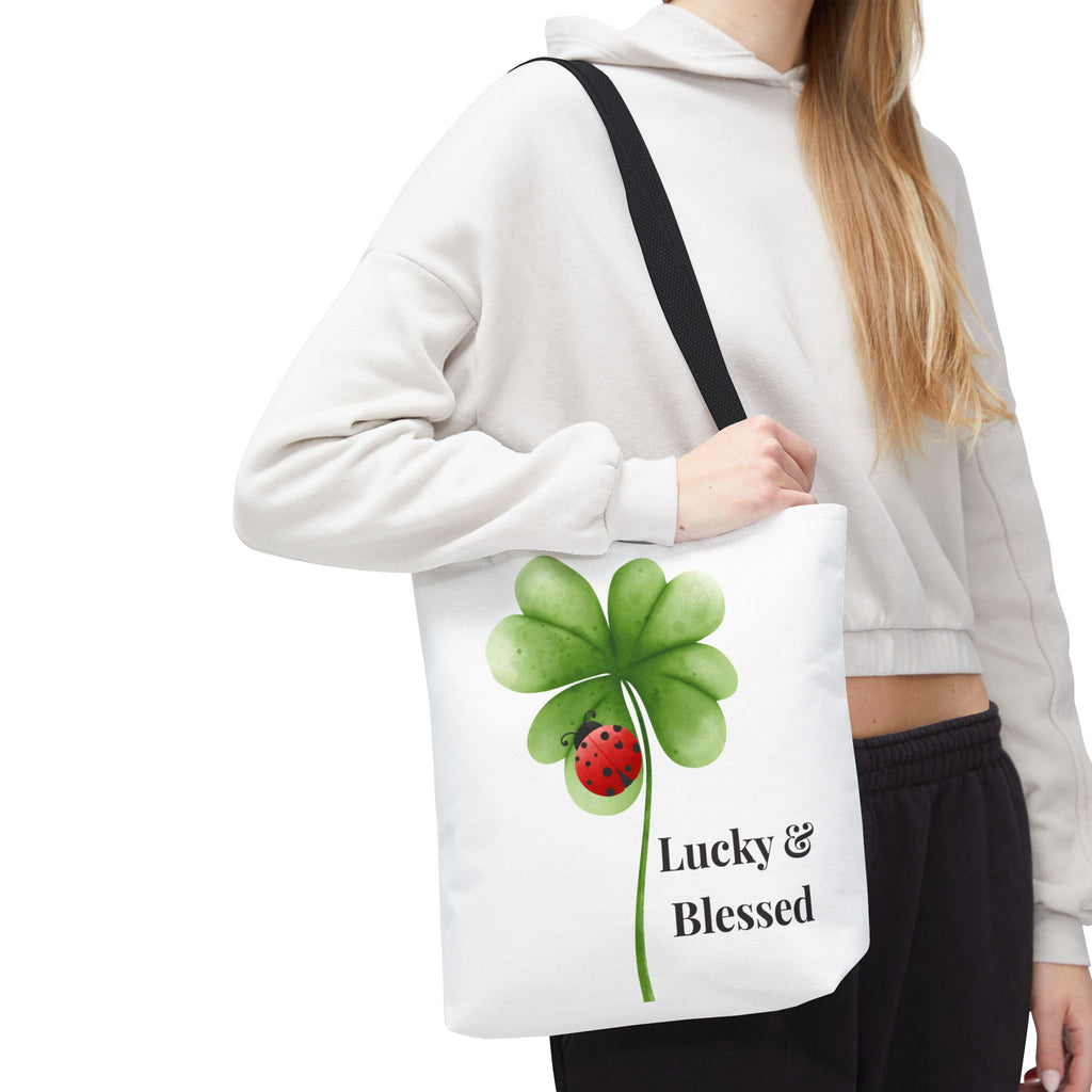 Person holding a tote bag with a four-leaf clover and ladybug design, text 'Lucky & Blessed', on a white background.