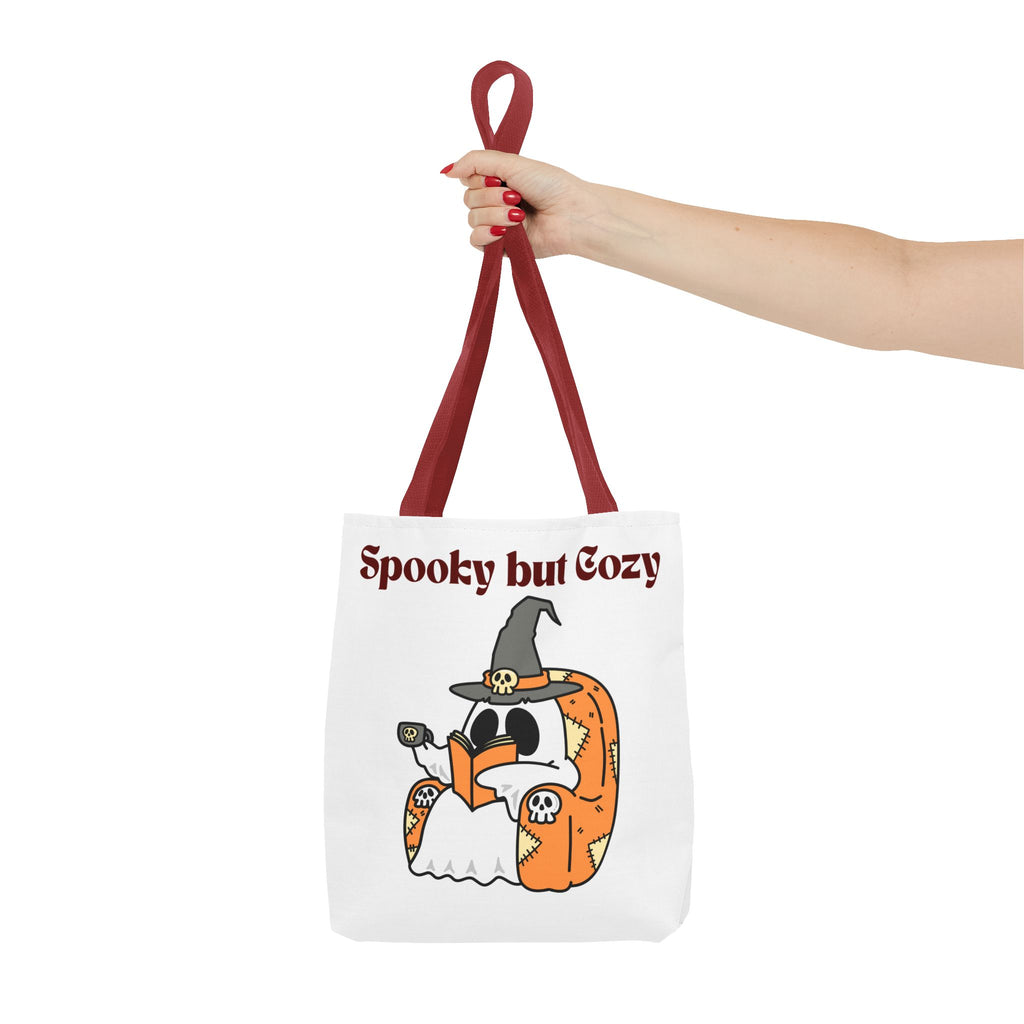 Tote bag with ghost and pumpkin design and 'Spooky but Cozy' text, held by a hand.