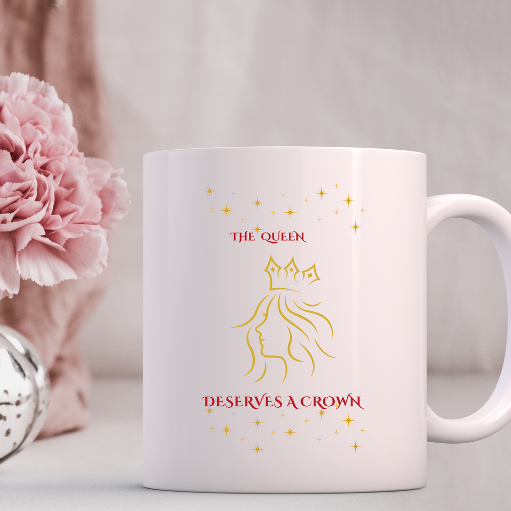 White mug with gold and red design featuring a woman's silhouette and 'The Queen Deserves a Crown' text, next to pink flowers.